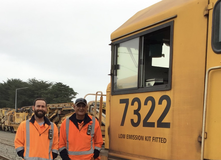 Two men stand next to the cab of a yellow locomotive, which reads '7322 LOW EMISSION KIT FITTED'